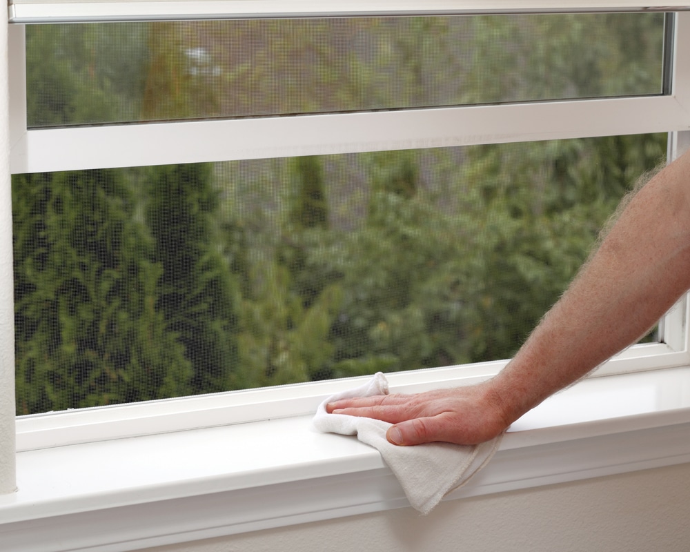 Hand using rag to clean below open window, pine trees outside